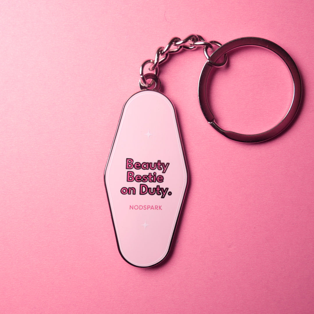 Pink motel-style keychain fob printed with "Beauty Bestie on Duty." displayed on a vibrant pink background.