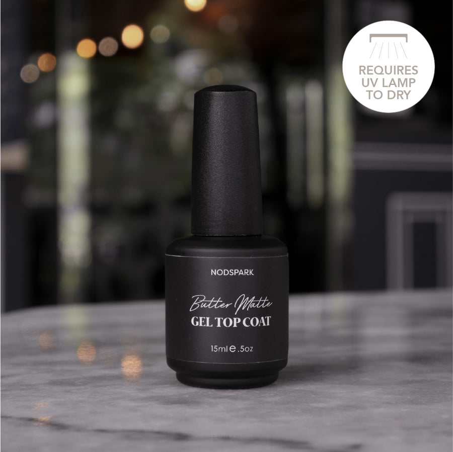 Black bottle of Nodspark Butter Matte Gel Top Coat shown upright on a smooth table with a warm, soft-focus background.