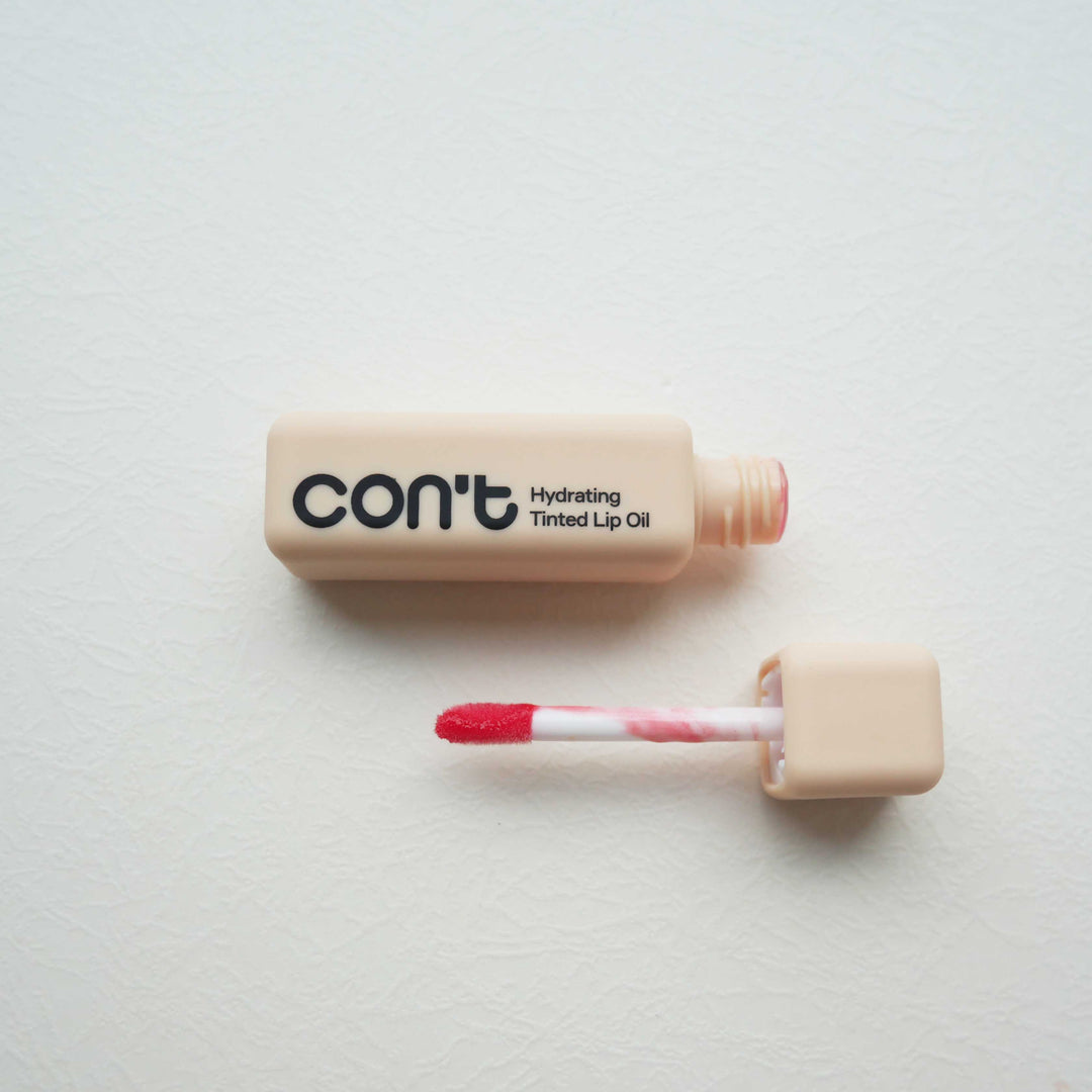 Square beige tube of Con't Hydrating Tinted Lip Oil in Coral shown open next to its coral-tinted applicator wand on a light neutral surface.