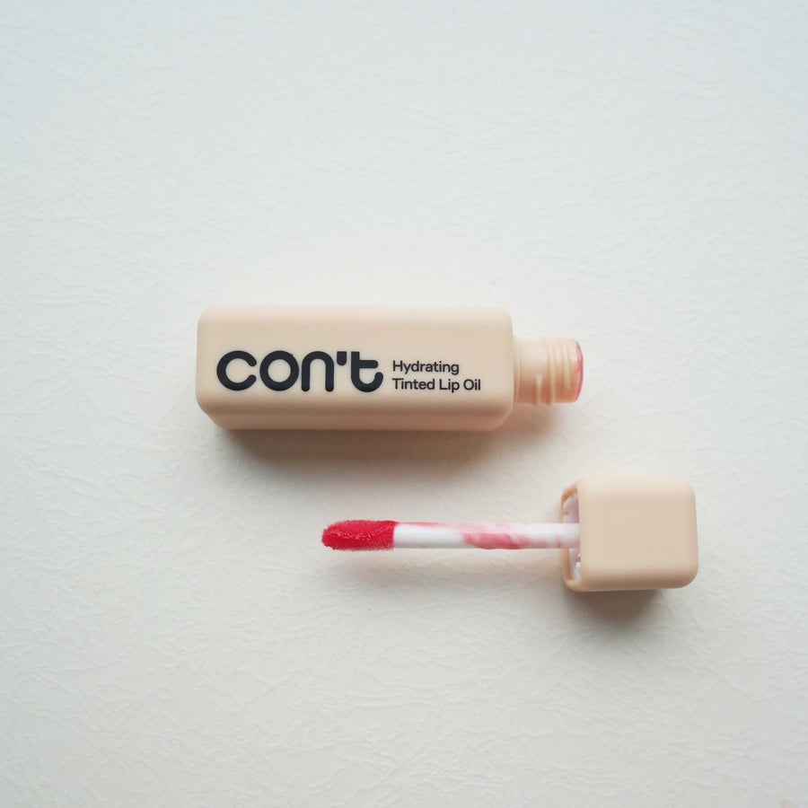 Square beige tube of Con't Hydrating Tinted Lip Oil in Coral shown open next to its coral-tinted applicator wand on a light neutral surface.