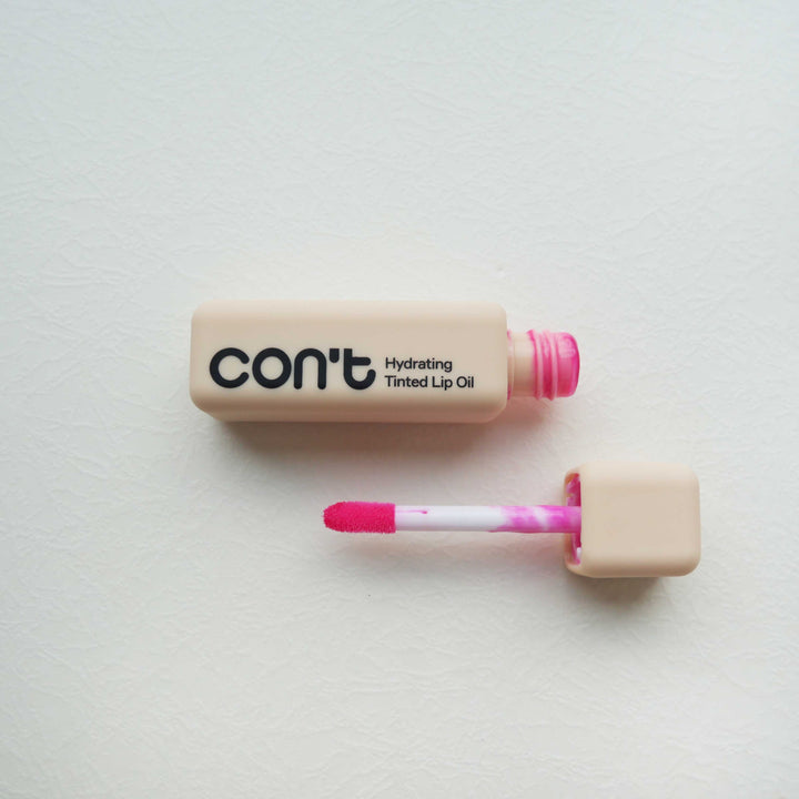 Square beige tube of Con't Hydrating Tinted Lip Oil in Rose with its pink-tinted applicator wand open beside it.