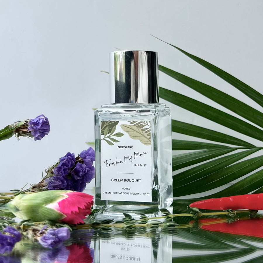 Clear spray bottle of Green Bouquet Hair Mist with a silver cap set among tropical green leaves and fresh flowers.