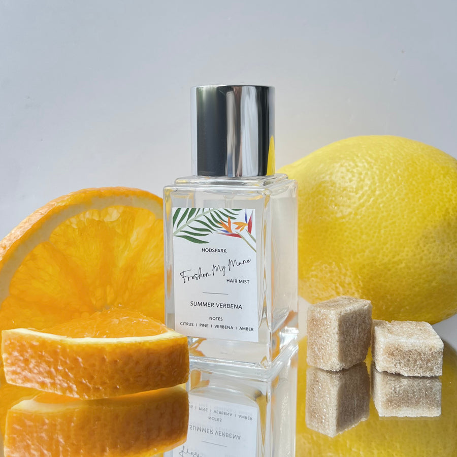 Clear spray bottle of Summer Verbena Hair Mist displayed with fresh citrus slices, a whole lemon, and sugar cubes.
