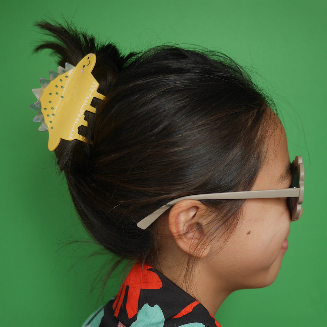 Side profile of a person wearing a yellow stegosaurus-shaped hair claw clip in their dark hair against a green background.