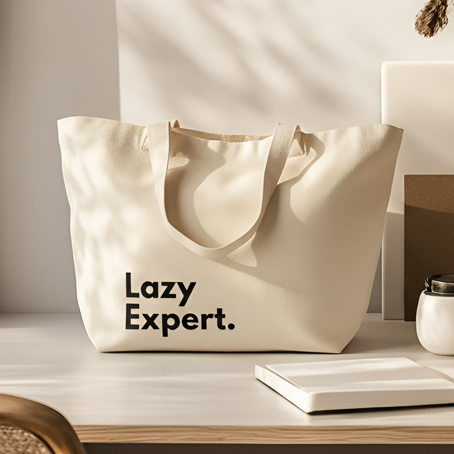 Off-white canvas tote bag printed with the words "Lazy Expert." resting on a bright wooden tabletop next to a notebook.