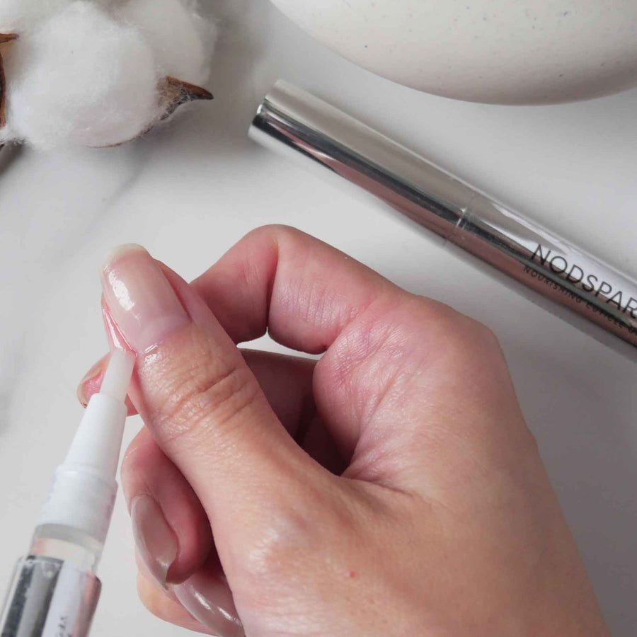 A hand applying cuticle oil around the nail using a slim metallic applicator pen with a precise tip.