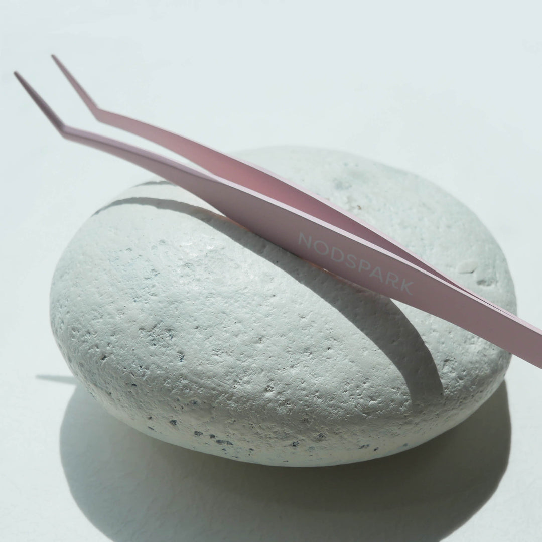 Light pink curved precision eyelash applicator tweezers resting horizontally on a textured white round surface.