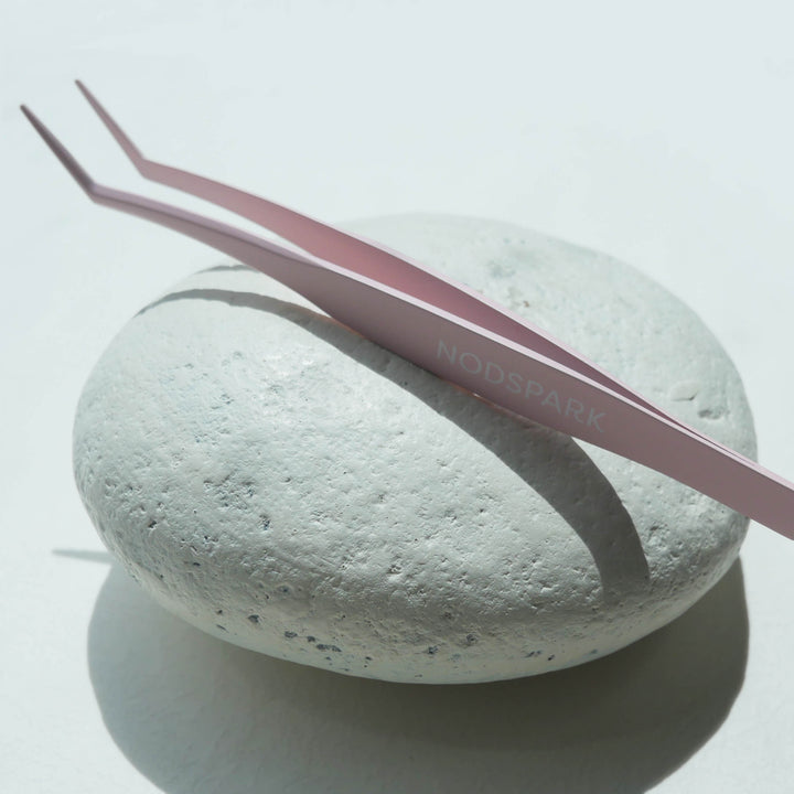 Light pink curved precision eyelash applicator tweezers resting horizontally on a textured white round surface.