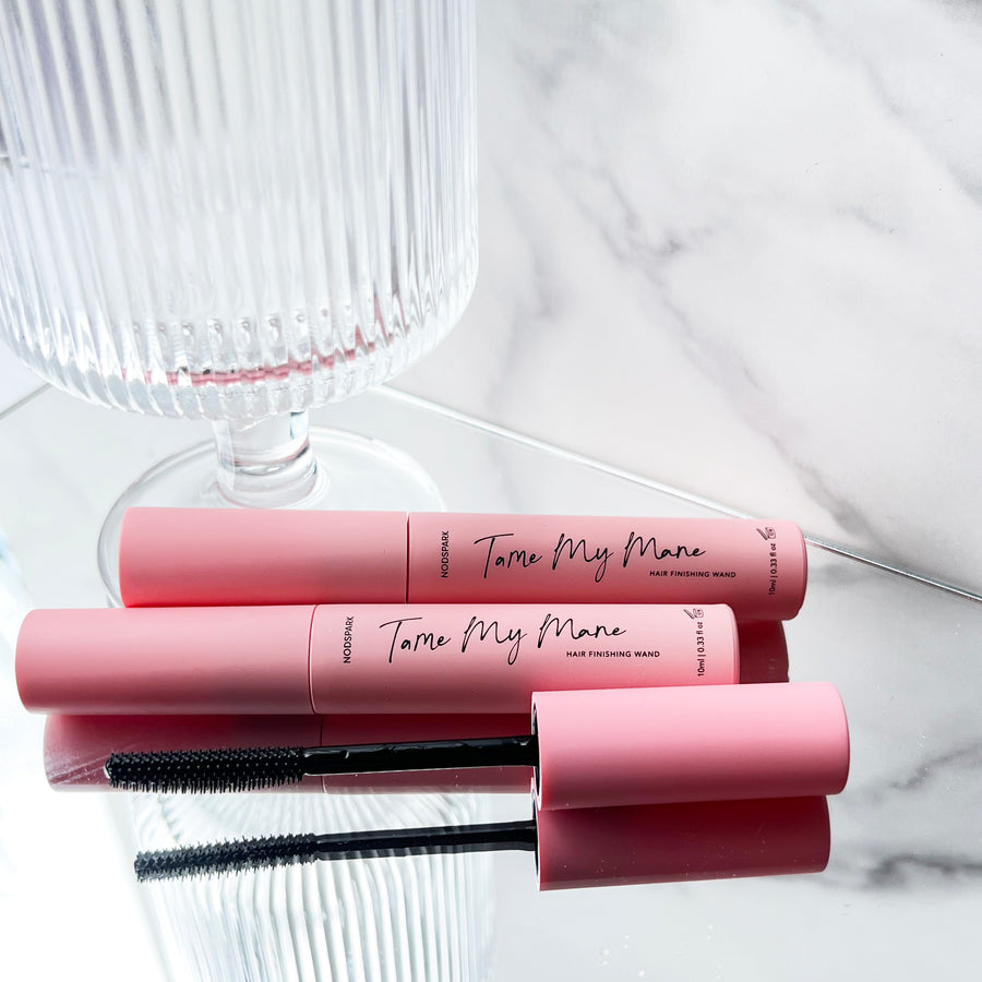 Three pink mascara-style tubes of Tame My Mane Hair Wand stacked alongside an open spoolie brush on a light background next to a glass cup.