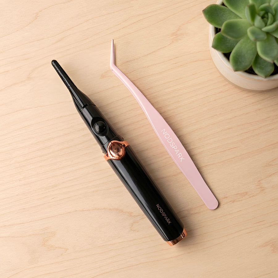 Top-down view of a black and rose-gold heated eyelash curler alongside pink lash tweezers, both featuring  NODSPARK logos, laid on a smooth light wood surface with a small green succulent plant on the right.