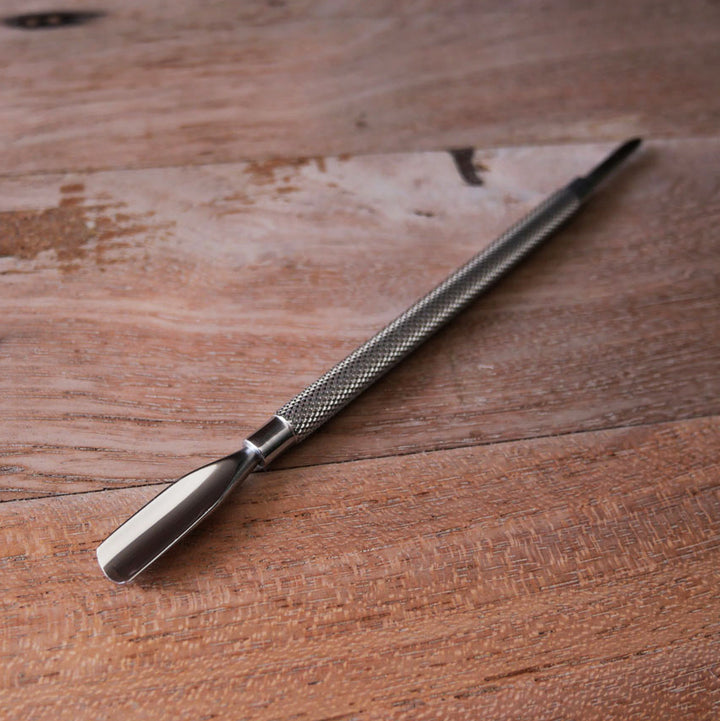 Dual-ended stainless steel cuticle pusher tool lying diagonally across a wooden surface.