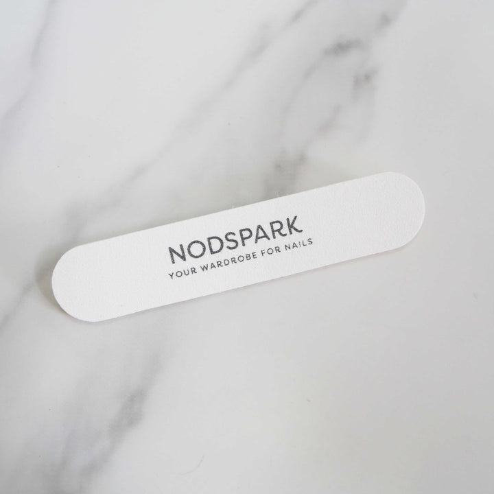 White disposable Nodspark nail file lying diagonally on a smooth marble surface.
