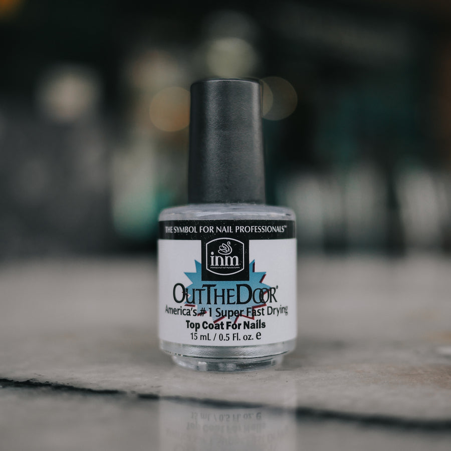 Clear glass bottle of INM Out The Door Top Coat with a black cap, displayed on a light polished surface.